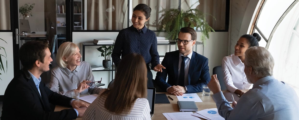 A diverse group of business professionals sit around a conference table while one woman stands and leads the discussion during a team meeting.